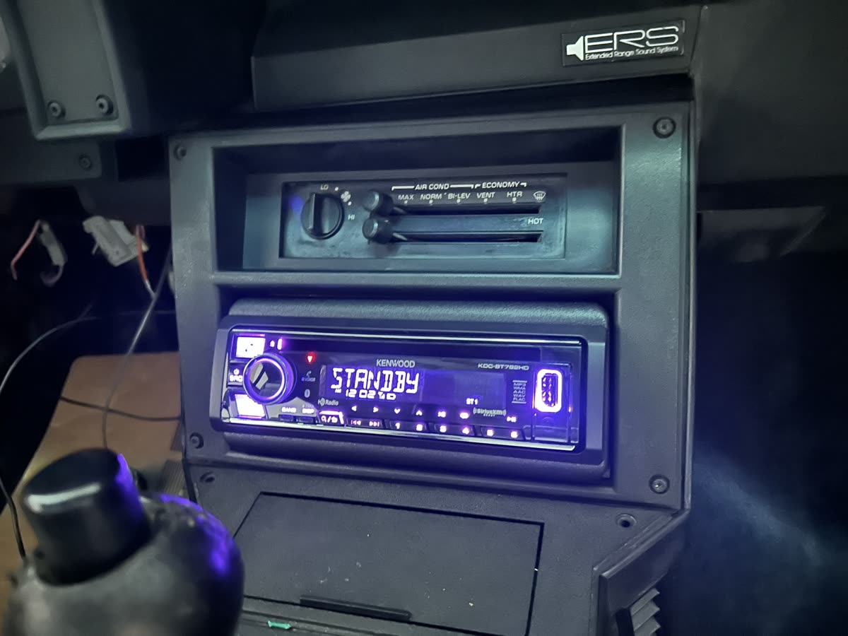 Kenwood single-DIN radio mounted in a dash kit with a storage pocket below